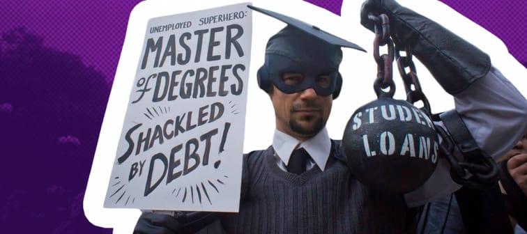 Man wearing a cap and gown holds a ball and chain and a sign advocating for cancelling student debt.