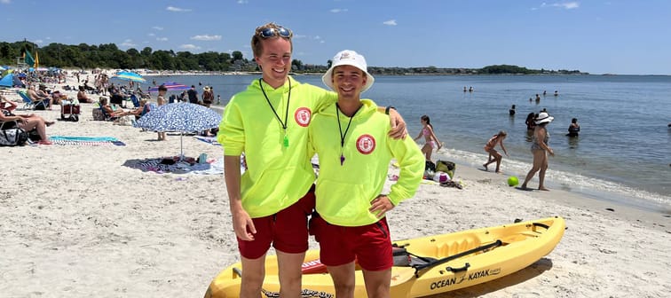 Ben Schies and Ryan Anderson, two young lifeguards, smile with arms around each other on the beach.