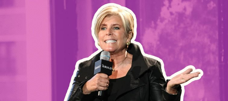 Suze Orman gestures with her hand, talking into a microphone on a stage.