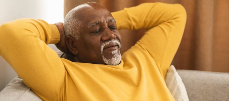 Senior Man Sleeping Holding Hands Behind Head Sitting On Sofa At Home.