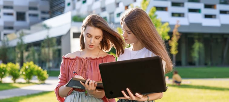 Two college students are standing near the university, looking at a computer, making notes in a notebook.