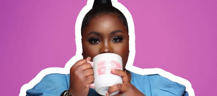 Dasha Kennedy poses for the camera taking a sip from a white coffee mug in front of a pink background.