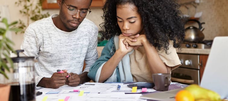 Young African couple doing paperwork together, sitting at kitchen table with lots of papers, calculator and laptop, looking frustrated. Dark-skinned family calculating domestic budget at home