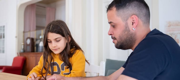 Father helps young girl with homework at kitchen table.