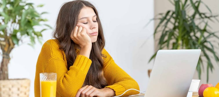 Young girl fell asleep in front of laptop. Cute woman is bored, tired or overworked. Cozy home environment. Pretty female in bright yellow jumper working on laptop at home