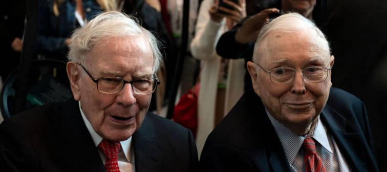 Charlie Munger and Warren Buffett sit side by side at a meeting, wearing suits and looking serious.