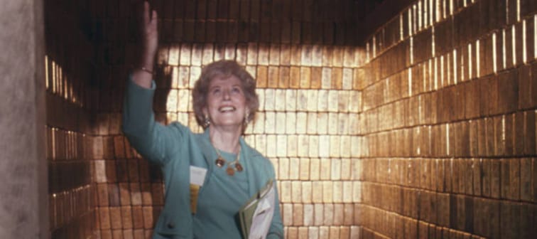 A woman stands in a small room lined with gold bar, holding her hand up and looking amazed.