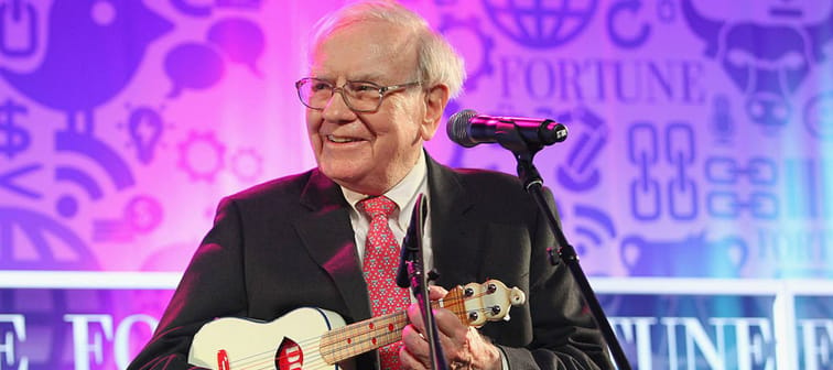 Warren Buffett on stage, looking to the side of the camera, holding a ukele and smiling.