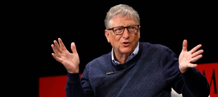 Bill Gates speaks on stage with his hands help up, in the middle of a thought.
