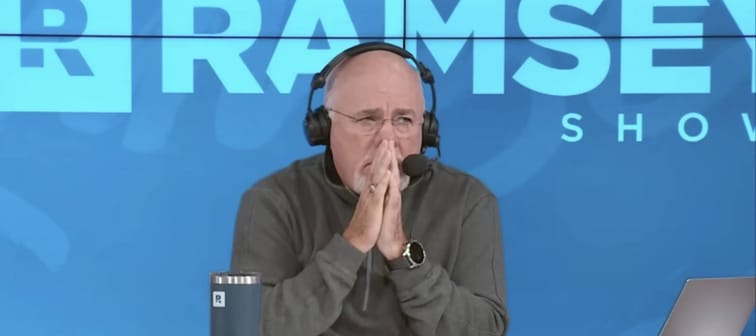 Dave Ramsey sits on set of his show with headphones on and his hands clasped in front of his mouth.