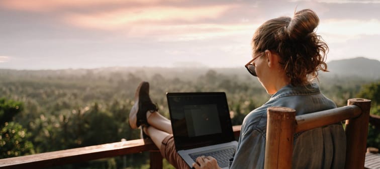 A woman using her computer with her feet up on a deck rail overlooking a wooded vista