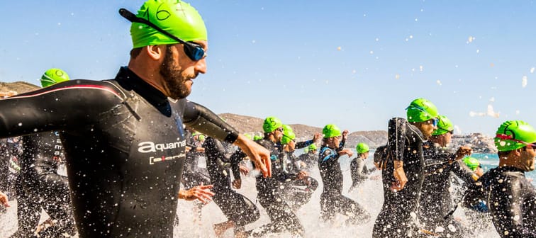 People in wetsuits and goggles run into a body of water.