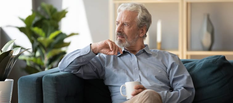 Thoughtful older man sitting on couch at home alone, holding cup of tea or coffee, feeling lonely and sad, unhappy depressed mature male looking into distance, thinking about problems