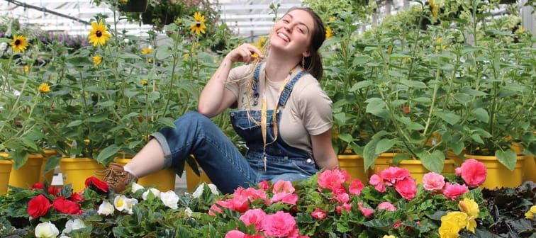 Juliet Del Rio, young woman smiling and facing camera surrounded by brightly coloured flowers in greenhouse