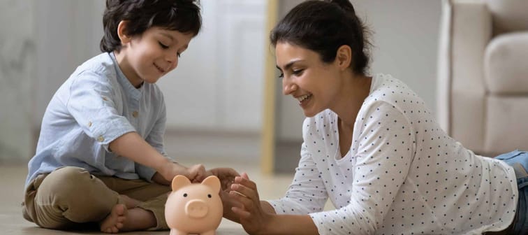Happy Indian kid and young mom saving money together, putting cash into ceramic piggy bank. Teaching child to invest money, planning future