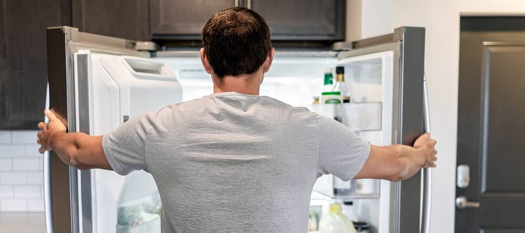 Back of hungry man opening fridge refrigerator doors domestic appliance searching for food inside with condiments and juice in modern kitchen