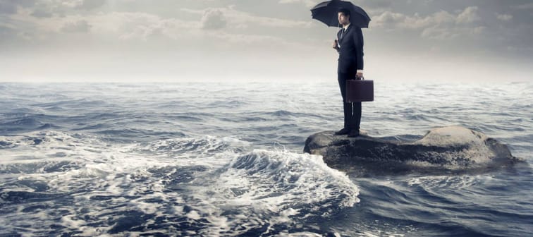 Businessman under an umbrella standing on a rock in the middle of a slight sea