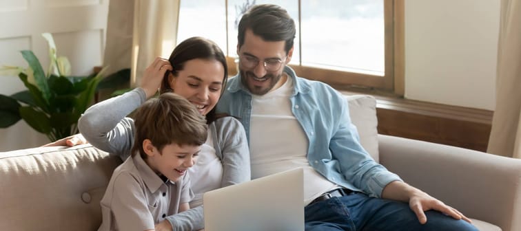 Smiling parents relax on sofa in living room with small 7s son child have fun browsing using modern laptop together