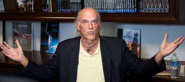 Jesse Ventura poses with his arms held out, about to say something in front of a desk with books on display.