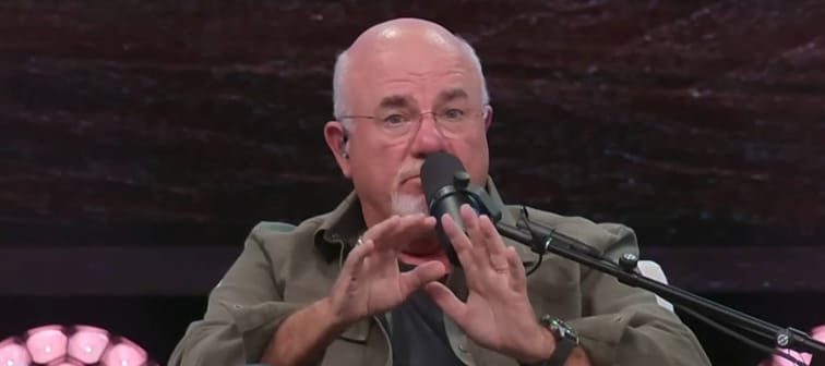 Dave Ramsey sits in a chair on a stage, talking into a microphone and making a surprised face.