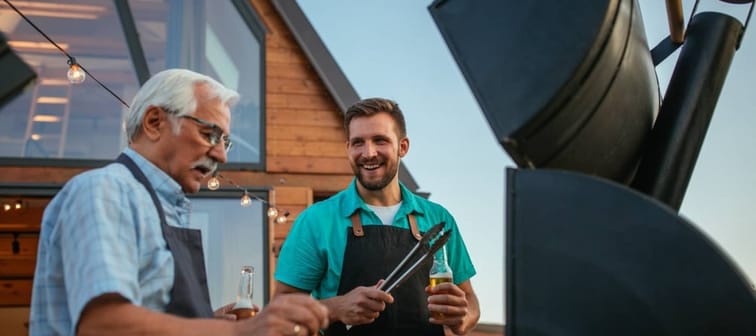 Father and son standing in front of barbecue, holding tongs and beers in their hands.