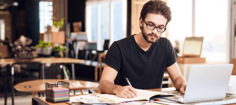 Freelancer bearded man in t-shirt taking notes at laptop sitting at desk.