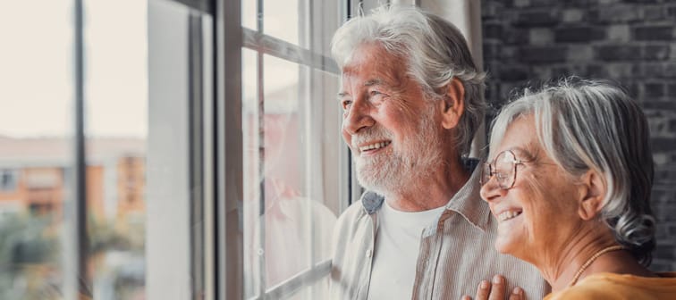 Happy bonding loving middle aged senior retired couple standing near window, looking in distance, recollecting good memories or planning common future