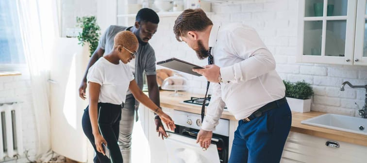 Professional bearded chubby man in formal outfit with clipboard inspecting oven together with attentive ethnic couple while standing at modern sunny apartment