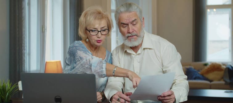 Senior aged couple checking accountancy and bills. Older retired couple holding computer and financial paper documents, checking domestic bills, discussing expenses together at home.