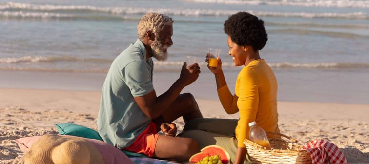 African american couple toasting juice while sitting with food on blanket against sea at sunset. retirement