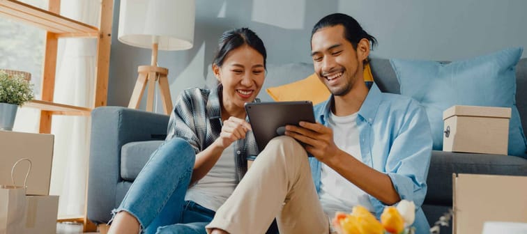 Happy asian  couple man and woman looking at tablet together seated on floor in front of couch