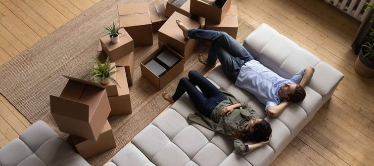 Top view happy couple tenants homeowners relaxing on couch on moving day in living room with cardboard boxes, man and woman resting after relocation in first own apartment, mortgage or rent
