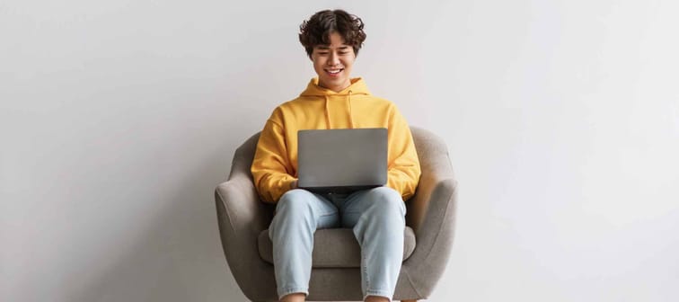 Happy young Asian male working online, sitting in armchair and using laptop