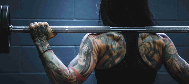 rear picture of tattoed woman's back at gym holding barbell with weights standing ready to do a squat to get strong