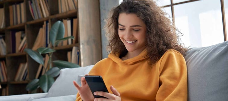Happy millennial hispanic teen girl checking social media holding smartphone at home.