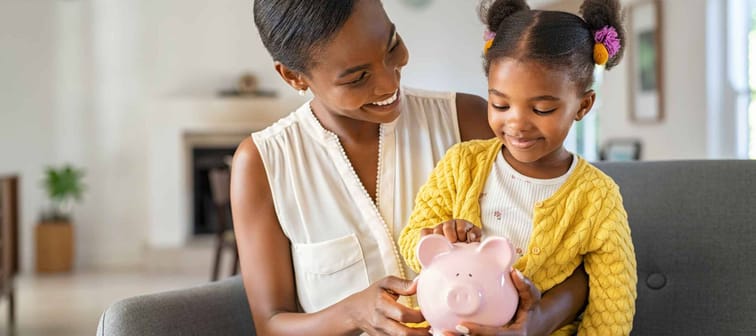 Smiling mature african american mother helping daughter sitting on lap putting money in piggy bank. Cute little black girl saving money by adding a coin in piggy bank with mother at home.