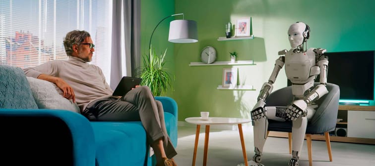 Full body middle aged man talking and using tablet to turn off futuristic android while sitting on sofa at home