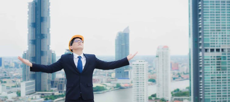 Happy Asian male engineer or businessman Wearing a yellow helmet is standing on the rooftop of building and a skyscraper over a cityscape