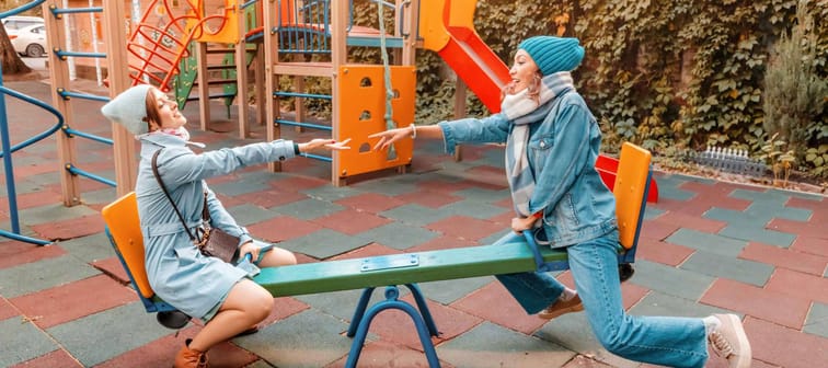 Two childish happy woman friends or teenage girl having fun on seesaw on a playground. Concept of psychology of the new generation and relations