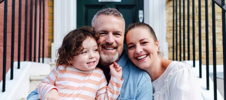 Happy family of three sit on front porch of house, smiling for camera.