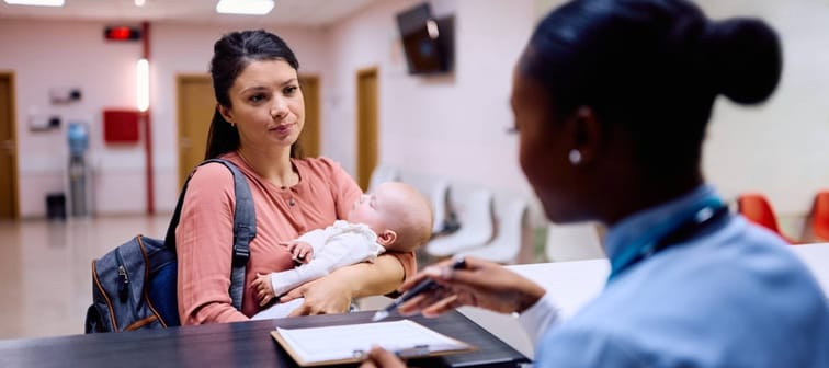 Mom holding baby while talking to receptionist at a doctor's office.