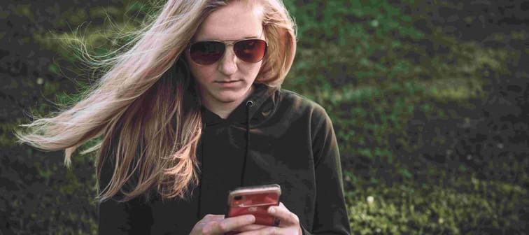 blonde woman standing in a field wearing glasses holding a cellphone wondering why