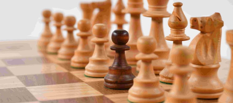 white chess pieces on a board lined up but one dark chess pawn stands among them as different from the rest