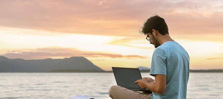 Young digital nomad man sitting on wooden pier at sea working on internet remotely at sunset - Traveling with a computer - Online dream job concept - Selective Focus