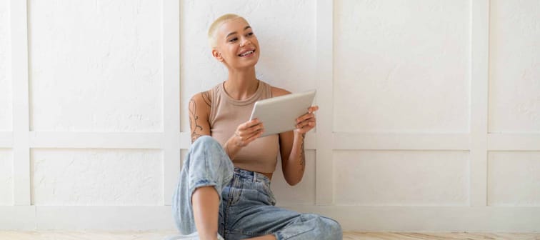 Smiling lady using digital tablet and looking aside at copy space, sitting on floor over white wall, free space. Happy woman posing with pad. Gadget lifestyle and online technology concept