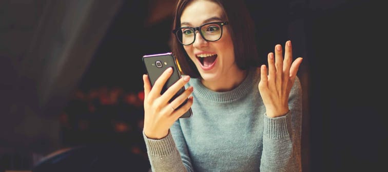 Surprised young female blogger chatting with best friends in social network on mobile phone while received shocking good news