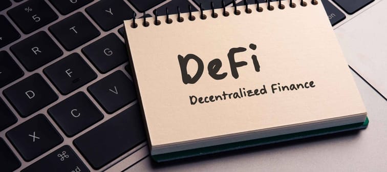 There is note book with the word DeFi (Decentralized Finance) on a laptop. It is an eye-catching image.