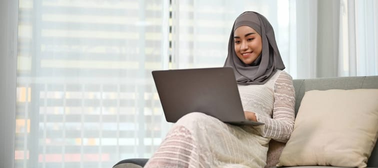 a young Muslim woman sitting on a couch with an open laptop