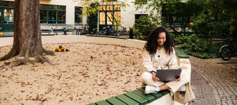 Smiling young african girl wearing autumn coat using laptop while sitting in lotus pose near sandbox