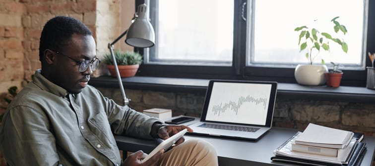 Horizontal medium shot of young Black man sitting at desk in office working with stock trading charts and graphs